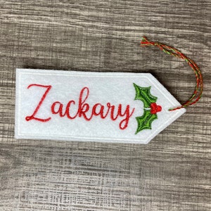 May include: White felt gift tag with the name "Zackary" embroidered in red script. A green holly and red berry design is on the right side. The tag has a red and green string for attaching to a gift.