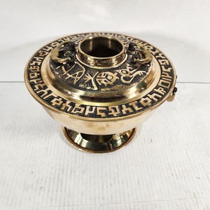 Könnte beinhalten: Ein Messing-Räuchergefäß mit rundem Sockel und schalenförmigem Körper. Der Deckel weist aufwendige Schnitzereien und eine zentrale Öffnung auf. Der Brenner hat ein dekoratives Band mit geätzten Symbolen am Rand. Das Gesamtdesign ist verziert und detailliert.