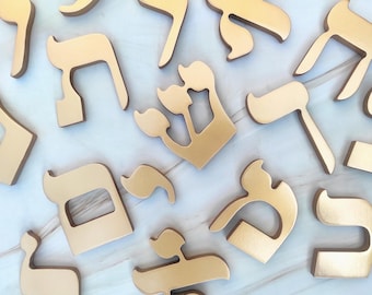May include: A collection of gold-colored Hebrew alphabet letters scattered across the frame. Each letter features a smooth, matte finish and distinct, stylized shapes representing various characters from the Hebrew script in a uniform gold tone.