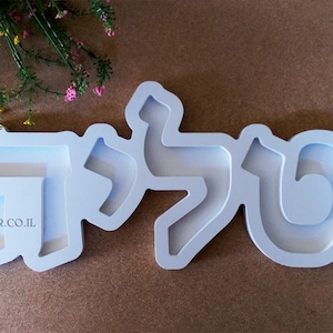 May include: White plastic mold for making a nameplate with the Hebrew letters "תל" in a modern font style. The mold is on a brown surface.