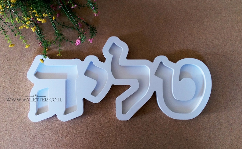 May include: White plastic mold for making a nameplate with the Hebrew letters "תל" in a modern font style. The mold is on a brown surface.