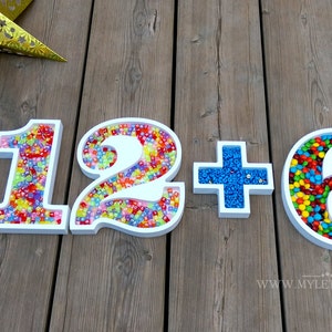 May include: White numbers 1, 2, and 6 filled with colorful candies, with a white plus sign between the numbers. The numbers are on a wooden surface.
