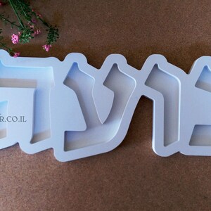 May include: White plastic mold for making a nameplate with the Hebrew word "שלום" (shalom) in raised letters. The mold is on a brown surface.