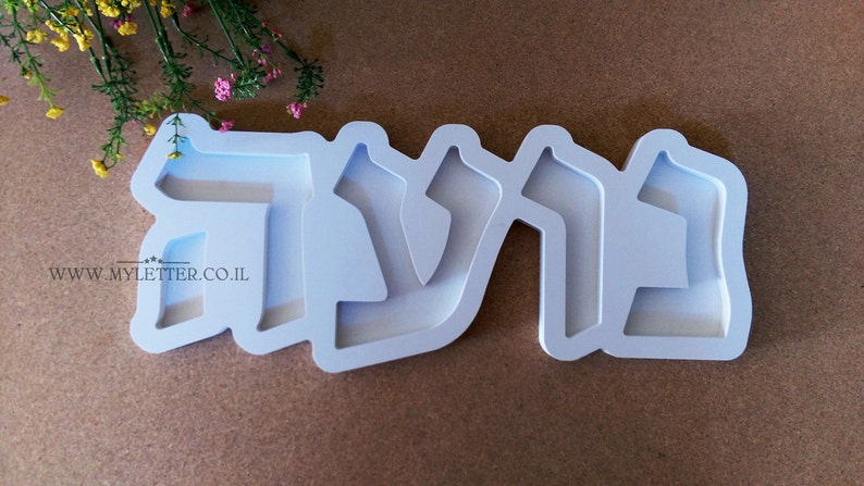 May include: White plastic mold for making a nameplate with the Hebrew word "שלום" (shalom) in raised letters. The mold is on a brown surface.