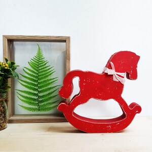 May include: Red wooden rocking horse with a pink ribbon. The horse is a decorative piece and is sitting on a wooden surface.