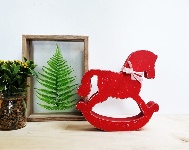 May include: Red wooden rocking horse with a pink ribbon. The horse is a decorative piece and is sitting on a wooden surface.