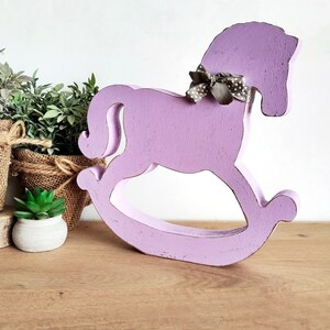 May include: A purple wooden rocking horse with a gray and white polka dot bow. The horse is on a wooden surface.