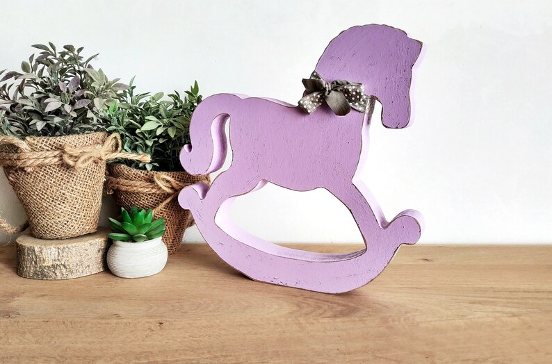 May include: A purple wooden rocking horse with a gray and white polka dot bow. The horse is on a wooden surface.