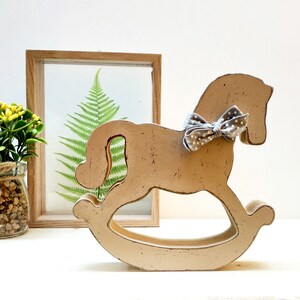 May include: A wooden rocking horse painted in a light beige color with a gray and white polka dot bow tied around its neck. The horse is sitting on a white surface.
