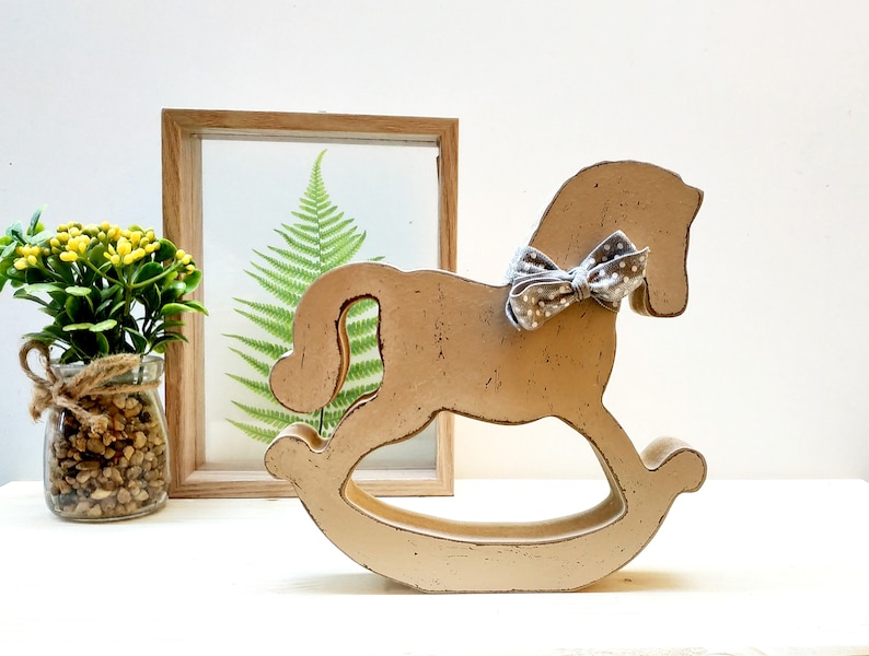 May include: A wooden rocking horse painted in a light beige color with a gray and white polka dot bow tied around its neck. The horse is sitting on a white surface.