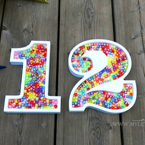 May include: White wooden numbers 1 and 2 filled with colorful alphabet beads. The numbers are on a wooden surface.