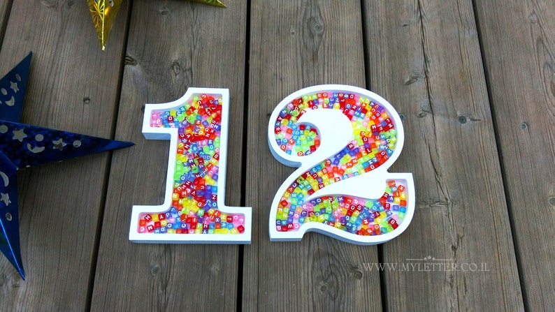 May include: White wooden numbers 1 and 2 filled with colorful alphabet beads. The numbers are on a wooden surface.