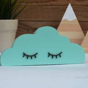 May include: A teal cloud-shaped wooden decoration with black eyelashes. Two wooden mountain decorations with white painted peaks are in the background. A white star and a potted plant are also visible.