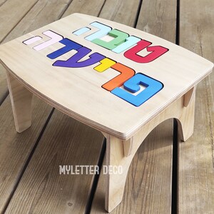Puede incluir: Taburete de madera con una parte superior de rompecabezas con letras hebreas de colores. Las letras son en tonos rosa, verde, rojo, naranja, azul y amarillo. El taburete tiene un acabado de madera natural y está diseñado para niños.