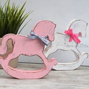 May include: Two wooden rocking horse figurines, one painted pink and the other white, with distressed paint finishes. Both horses have a bow tied around their necks, one gray and one pink with white polka dots. The pink horse is in the foreground and the white horse is in the background.