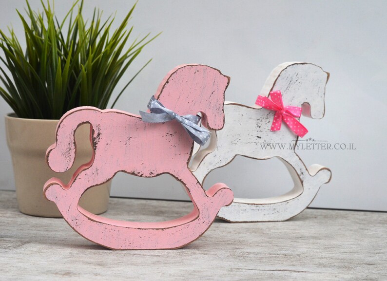 May include: Two wooden rocking horse figurines, one painted pink and the other white, with distressed paint finishes. Both horses have a bow tied around their necks, one gray and one pink with white polka dots. The pink horse is in the foreground and the white horse is in the background.