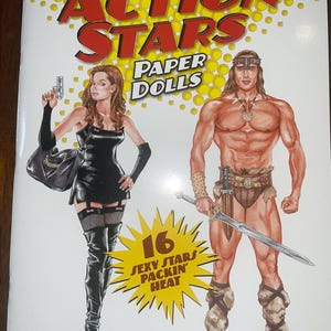 May include: A paperback book titled "Action Stars Paper Dolls" featuring illustrations of a woman holding a gun and a muscular man with a sword. The cover includes the text "16 Sexy Stars Packin' Heat" and the author's name, Bruce Patrick Jones.