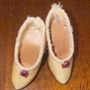 May include: A pair of miniature, cream-colored shoes with a pointed toe and a small, red jewel on the front. The shoes have a soft, fuzzy trim around the opening and a tan insole. These doll shoes are likely vintage.