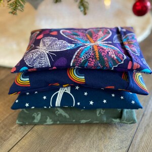 May include: Four fabric-covered heat packs stacked on top of each other. The top pack has a butterfly design in shades of pink, purple, and white. The second pack has a rainbow design on a navy blue background. The third pack has a white star pattern on a navy blue background. The bottom pack is a solid green color.