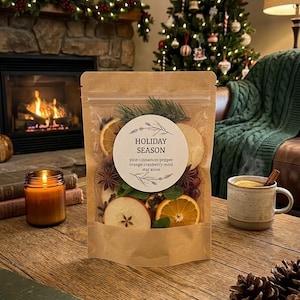 May include: A brown stand-up pouch containing dried fruit, star anise, and pine needles. A circular label reads HOLIDAY SEASON with the text pine-cinnamon-pepper, orange-cranberry-mint, star anise. The pouch is on a wooden table.