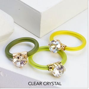 Op de afbeelding: Drie ringen met heldere kristallen stenen in goudkleurige metalen zettingen op flexibele siliconen banden. Twee banden zijn felgeel en één is olijfgroen. De tekst CLEAR CRYSTAL en CLEAR CRYSTAL | YELLOW SILICONE is zichtbaar.