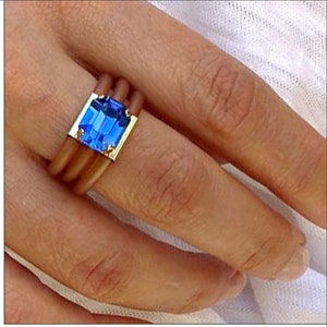 Op de afbeelding: Een ring met een rechthoekige, gefacetteerde blauwe edelsteen in een goudkleurige metalen zetting. De band bestaat uit drie gladde, beige, afgeronde parallelle strengen die om de vinger lopen voor een gelaagd effect.