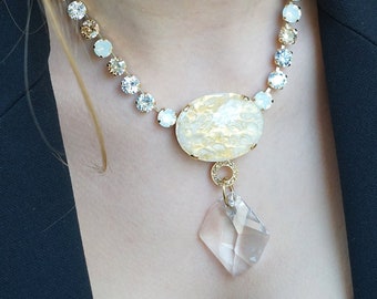 May include: A gold necklace with a white and gold oval pendant and a clear crystal drop. The necklace is made of small, round crystals in shades of blue, white, and gold.