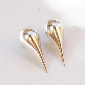 Op de afbeelding: Een paar zilveren oorbellen in de vorm van een traan met een puntige punt. De oorbellen liggen op een wit oppervlak. ANTIQUE SILVER staat in de rechterbenedenhoek van de afbeelding.