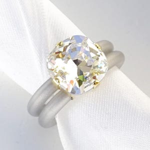以下が含まれることがあります： 輝くゴールドトーンの金属製プロングセッティングに、大きな透明のクッションカットストーンをあしらったリング。バンドはマットなグレーの二重リングで構成されています。隅には「CLEAR | SHINY GOLD」という文字が印字されています。