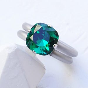 Op de afbeelding: Een ring met een vierkant geslepen smaragdgroen kristal in een gladde, mat zilverkleurige band. De tekst EMERALD | SHINY SILVER staat in de rechterbenedenhoek gedrukt. De ring is geplaatst tegen een witte, getextureerde ondergrond.