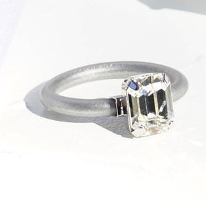 Op de afbeelding: Een ring met een matte zilverkleurige band en een enkele rechthoekige heldere gefacetteerde steen in een zilverkleurige metalen zetting. De tekst CLEAR | SILVER staat in de rechterbenedenhoek van de afbeelding.