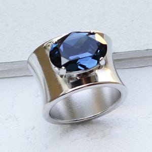 May include: A wide band ring featuring a large, faceted, oval-shaped navy blue stone set in a polished, shiny silver-tone metal setting. The text NAVY BLUE | SHINY SILVER is printed in the bottom right corner of the image.