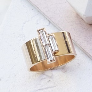 Puede incluir: Un anillo de oro rosa con un dise&ntilde;o de cristal transparente. El anillo es ancho y tiene una banda plana. El cristal est&aacute; engastado en una l&iacute;nea vertical y est&aacute; formado por tres piedras de forma baguette.