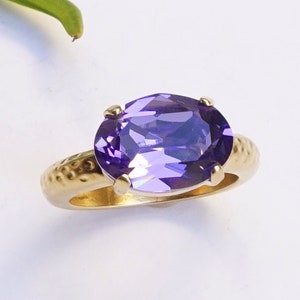 May include: A gold ring featuring a large, oval-cut purple gemstone set in four prongs. The band of the ring has a textured, hammered finish, providing a distinct contrast to the smooth, faceted surface of the vibrant purple stone.