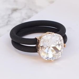 Puede incluir: Un anillo con una banda doble de silicona negra y un cristal transparente grande y facetado en un engaste de metal color oro rosa. El texto "CLEAR CRYSTAL | ROSE GOLD" está impreso en la esquina inferior derecha de la imagen.