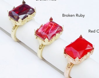 May include: Three gold rings with different red gemstone settings. The rings are arranged on a white surface. The gemstones are labelled "Broken Red", "Broken Ruby", and "Red Opaque".