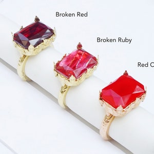 Op de afbeelding: Drie gouden ringen met verschillende rode edelsteen bezettingen. De ringen zijn gerangschikt op een wit oppervlak. De edelstenen zijn gelabeld "Broken Red", "Broken Ruby" en "Red Opaque".