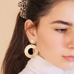 May include: Gold hoop earrings with a smaller circle dangling from the bottom. The earrings are worn by a person with long brown hair.