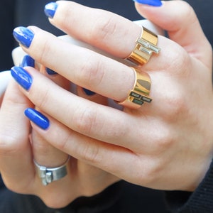 Puede incluir: Dos anillos de oro con piedras rectangulares en una mano con esmalte de u&ntilde;as azul.