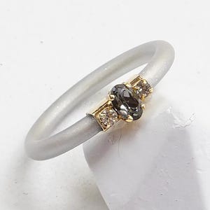 Op de afbeelding: Een grijze siliconen ring met een gouden band en drie stenen. De middelste steen is ovaalvormig en donkergrijs, terwijl de twee kleinere stenen helder zijn.
