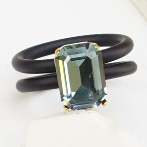 May include: A black silicone ring with a green emerald-cut gemstone. The ring is double-banded and has a gold setting.