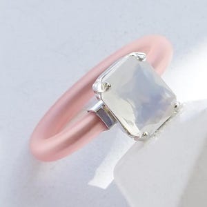 Op de afbeelding: Een ring met een gladde, matte pastelroze band en een rechthoekige, gefacetteerde melkwitte steen in een zilverkleurige metalen zetting. De tekst MILKEY WHITE | SILVER staat gedrukt in de rechterbenedenhoek van de afbeelding.