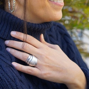 May include: A silver ring with a wavy design. The ring is worn on the index finger of a person wearing a blue sweater.