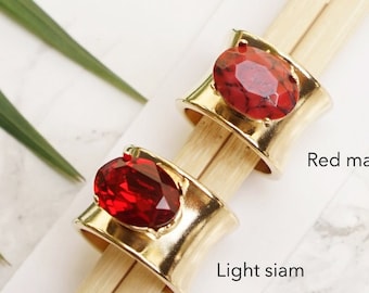 May include: Two gold-toned rings with wide bands, each featuring a large oval-shaped stone. One ring has a red marble stone labelled Red marble, and the other ring has a faceted red stone labelled Light siam. The rings are displayed on wooden sticks.