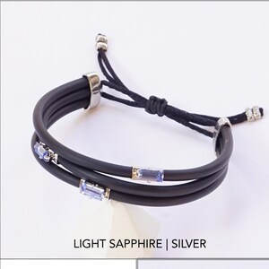 May include: Black adjustable bracelet with three strands of black cord and a silver clasp. The bracelet has three light blue gemstone accents. "LIGHT SAPPHIRE | SILVER" is written below the bracelet.