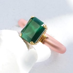 Op de afbeelding: Een ring met een rechthoekige smaragdgroene steen gezet in geelgoud, gemonteerd op een gladde, mat afgewerkte lichtroze band. De tekst EMERALD OPAQUE | YELLOW GOLD staat onderaan de afbeelding en beschrijft de steen en het metaal.
