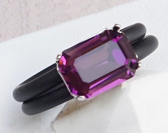 Op de afbeelding: Een zwarte rubberen ring met een paarse edelsteen in een zilveren zetting. De ring ligt op een wit oppervlak. AMETHYST staat onder de ring.