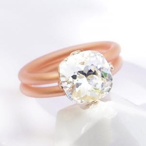 Op de afbeelding: Een ring met een grote, gefacetteerde heldere kristal op een dubbele ringband in roségoudkleur. De tekst CLEAR CRYSTAL | ROSE GOLD staat rechtsonder in de afbeelding. De ring is geplaatst op een wit geometrisch object.