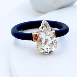 以下が含まれることがあります： 黒いリングに、透明な涙型の宝石が付いています。宝石は金色の金属にセットされています。リングは白い表面にあります。「CLEAR | BLACK」というテキストが画像の下部にあります。
