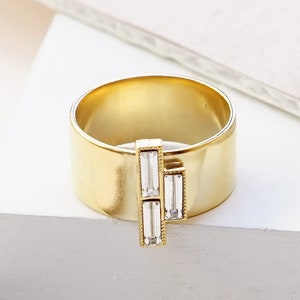 Puede incluir: Un anillo de oro mate con tres piedras transparentes de forma baguette. CLEAR | MATTE GOLD.